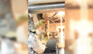 Ein Teammitglied der Raesch Quarz (Germany) GmbH, einem teilnehmenden Unternehmen bei der INDUSTRIE INTOUCH 2022, arbeitet in einem Schutzanzug an einem Quarzglasrohr. Im Hintergrund fliegen Funken.
