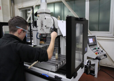 Das Bild zeigt eine Person, die an einer Standbohrmaschine in einer Fabrikhalle von FFT Produktionssysteme GmbH & Co. KG arbeitet. Die Person trägt eine Sicherheitsbrille.