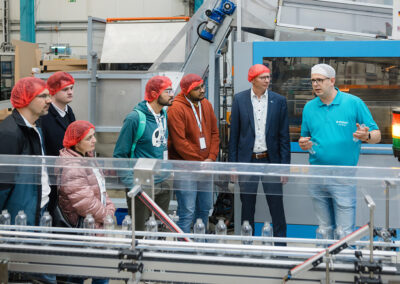 Ein Mitarbeiter der Firma EPROPLAST erklärt Besuchern mit roten Haarnetzen den Prozess der Flaschenherstellung an einer Maschine in einer Produktionshalle.