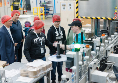 Ein Mitarbeiter der Firma EPROPLAST erklärt Besuchern mit roten Haarnetzen den Prozess der Flaschenherstellung an einer Maschine in einer Produktionshalle.